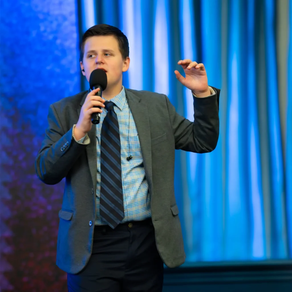 Man in a suit speaking into a microphone in front of blue curtains