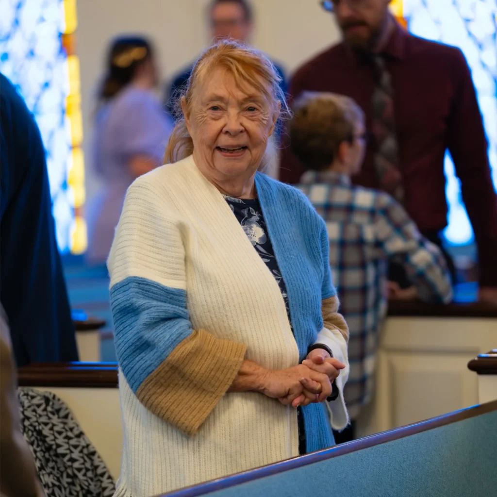 Older woman smiling while standing beside a church pew