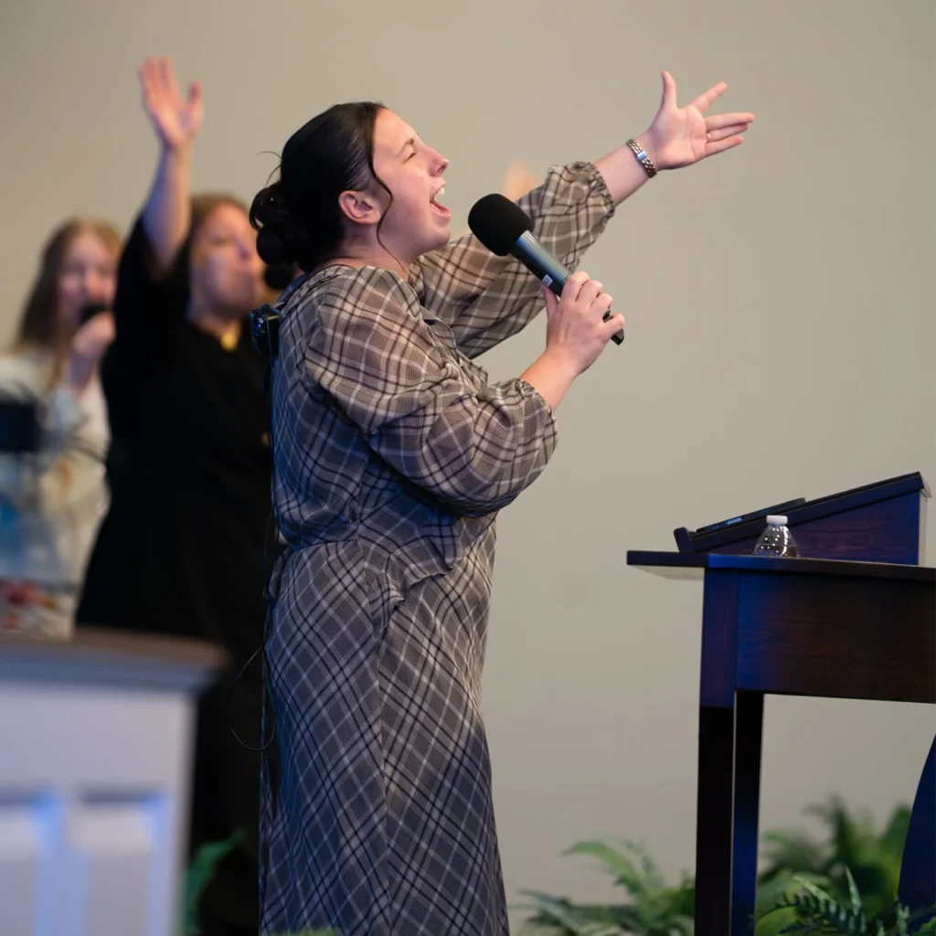 Woman singing into a microphone with one hand raised beside the pulpit